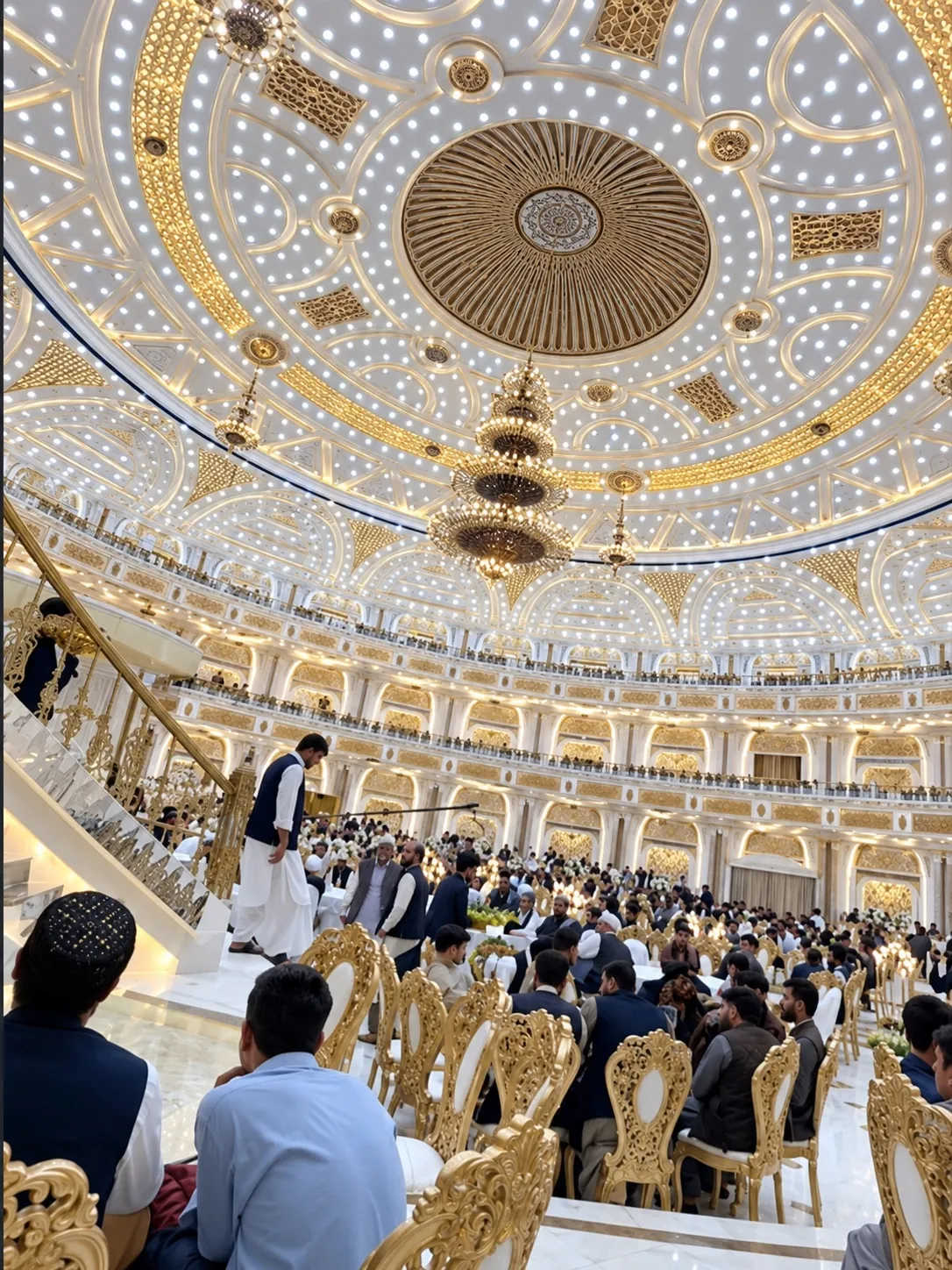 Interior of a large wedding hall with a gold domed ceiling and chandeliers above seated guests